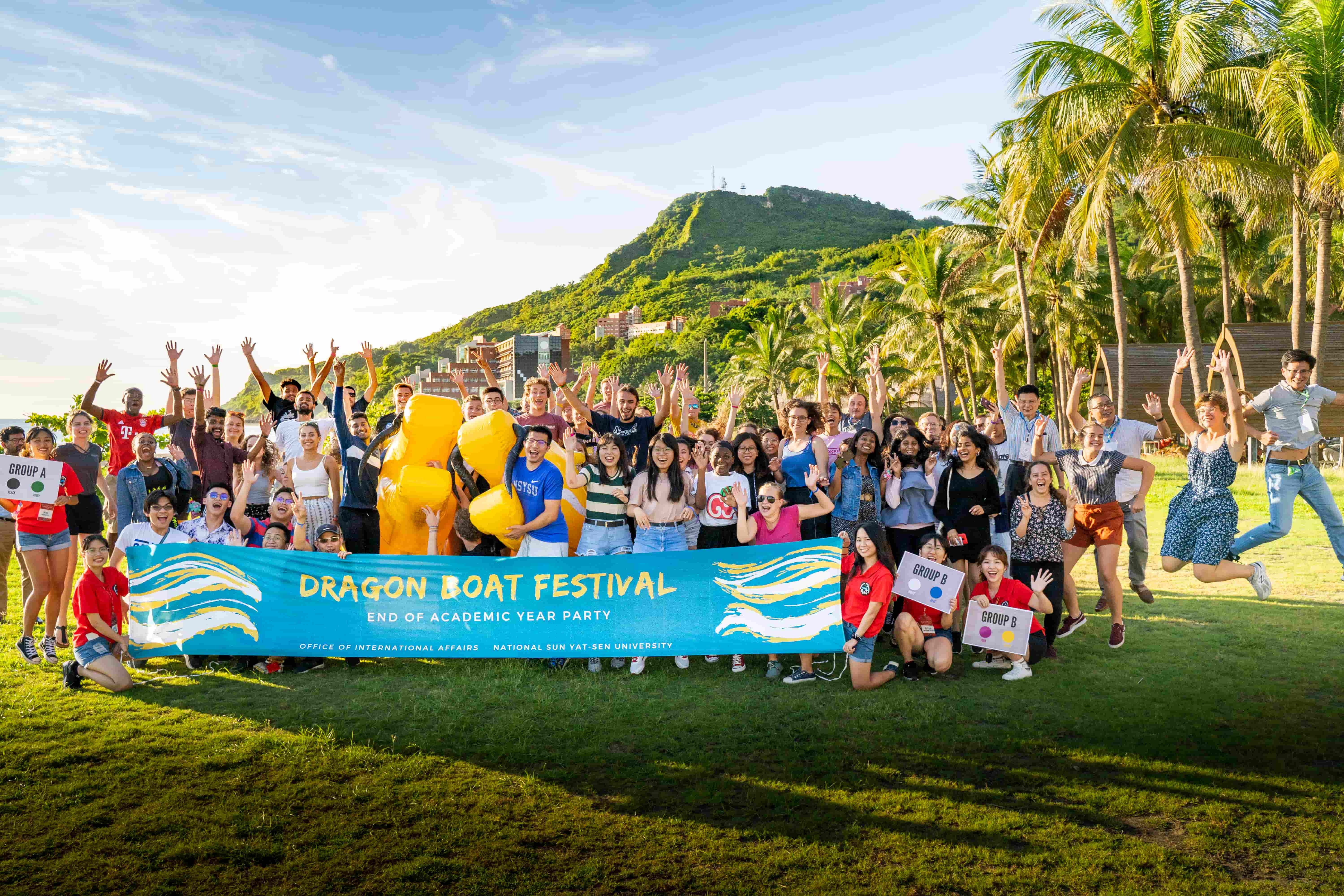 International students posing together outdoors on the NSYSU campus, with mountains, campus buildings and palm trees in the background.