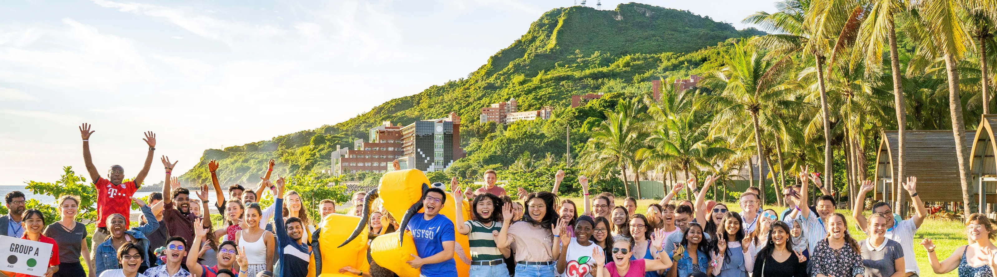 International students posing together outdoors on the NSYSU campus, with mountains, campus buildings and palm trees in the background.
