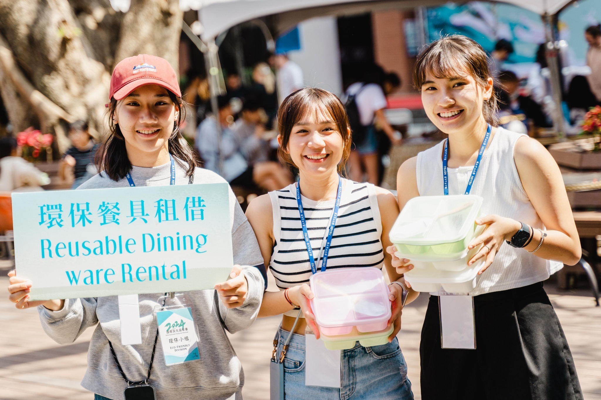 Three international students promote reusable dining ware rental at an outdoor cultural festival.